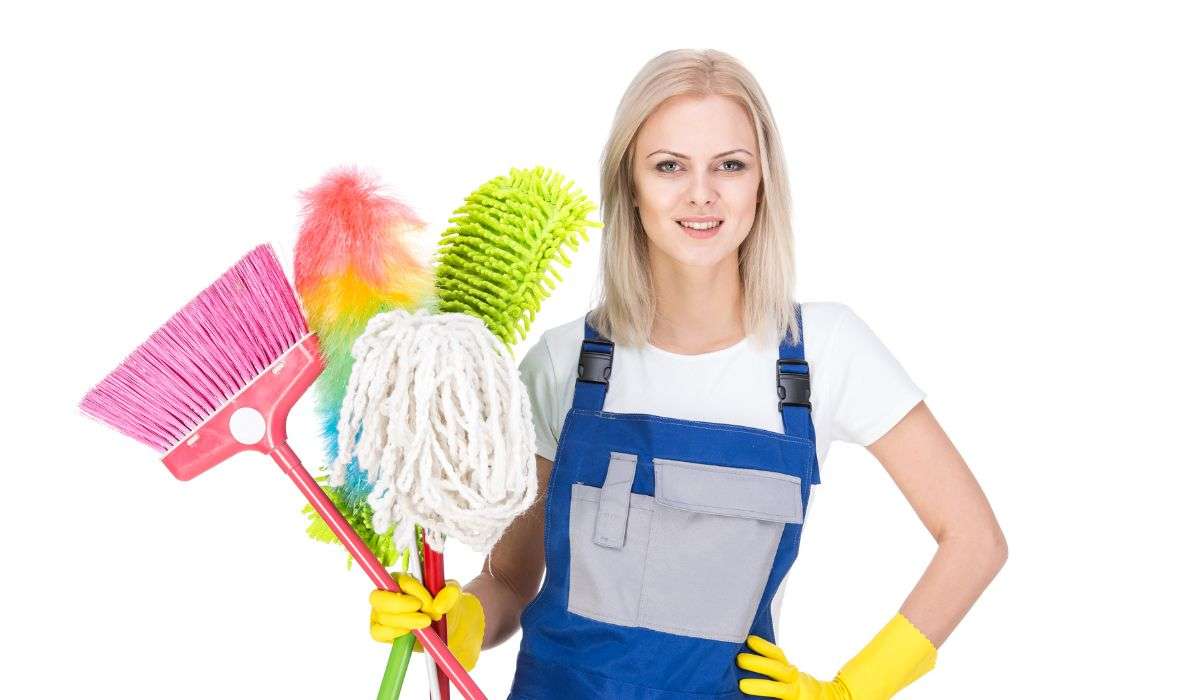 Blonde maid in blue uniform holding mop, broom, and duster on white background.