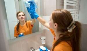 Maid cleaning a bathroom mirror with a cloth and spray bottle during residential cleaning service.