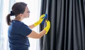 Woman cleaning curtains in a well-lit room using gloves and a cloth.